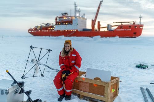 Friederike Krüger auf einer Eisscholle im roten, warmen Schneeanzug, im Hintergrund ist das Begleitschiff "Akademik Fedorov" zu sehen.©  Quelle: Mario Hoppmann